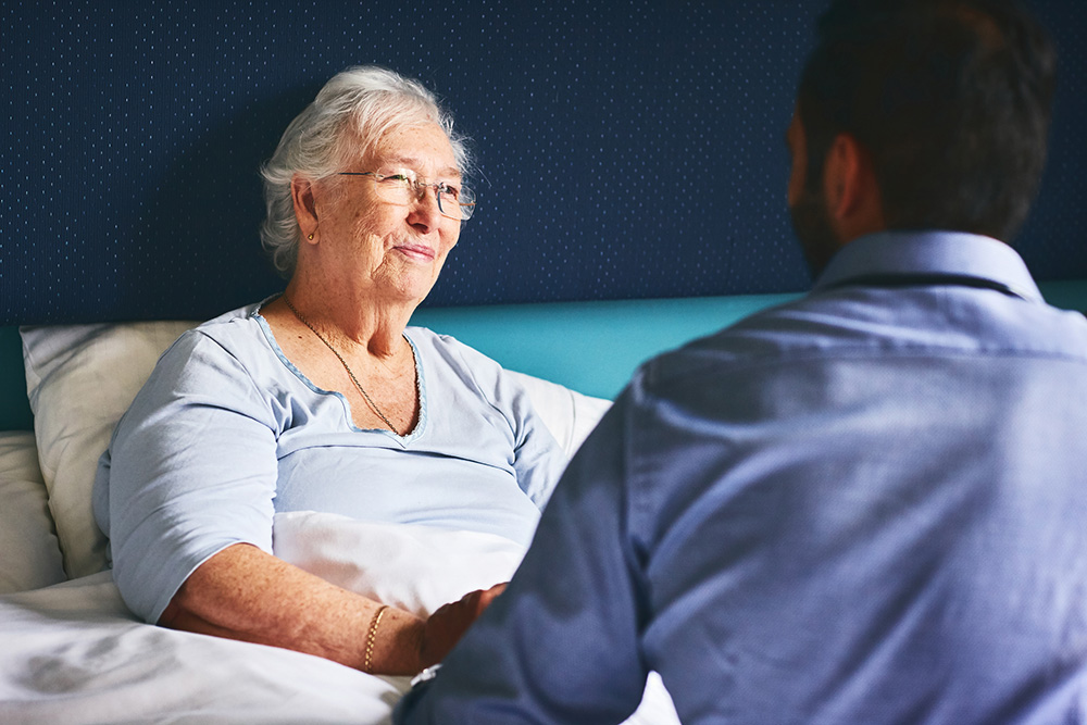 Happy senior female patient lying on bed with doctor comforting her at nursing home